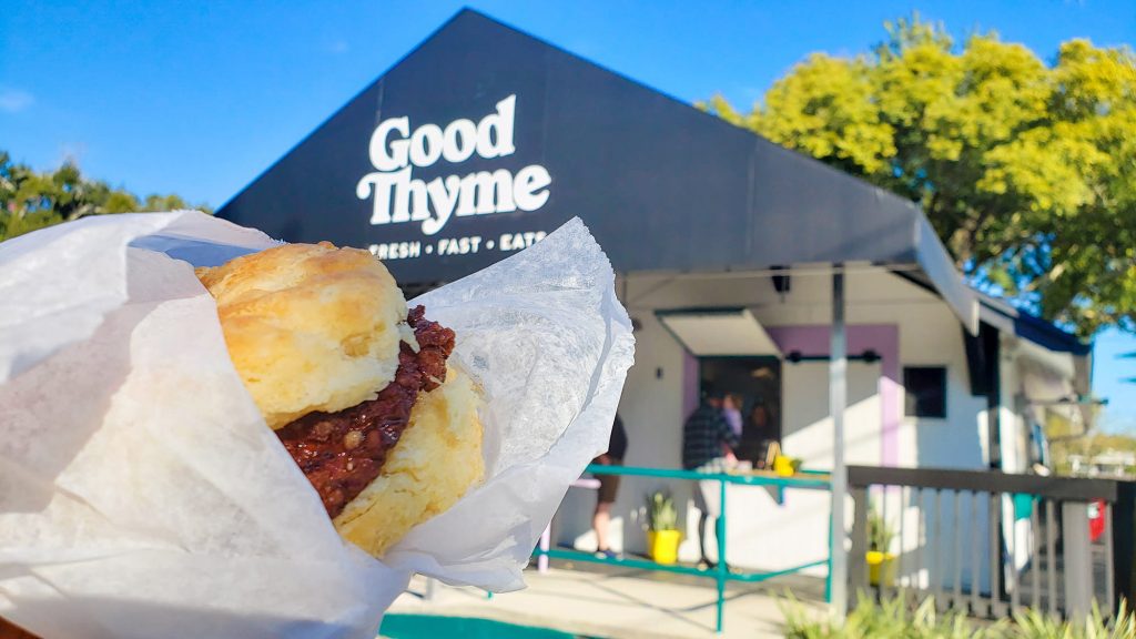 chicken biscuit being held up in front of Good Thyme LKLD. People ordering food in background.